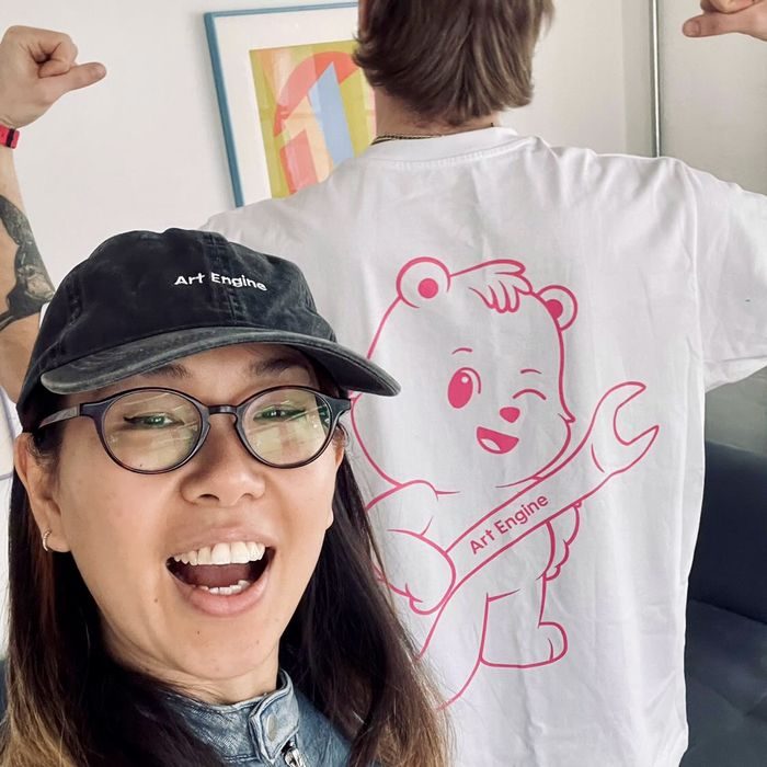 A smiling woman in an Art Engine cap, the winking bear on a tee shirt behind her